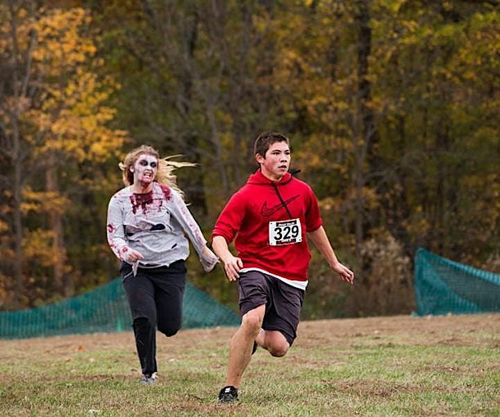 A runner sprints across a park field with a zombie in torn, bloodied clothes chasing close behind
